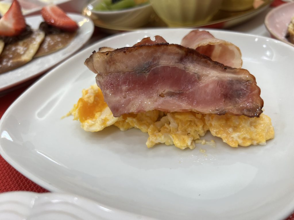 A white plate with cheddar cheesy scrambled eggs and a slice of bacon at MB Cakes Brunch & Coffee Photos & Review
