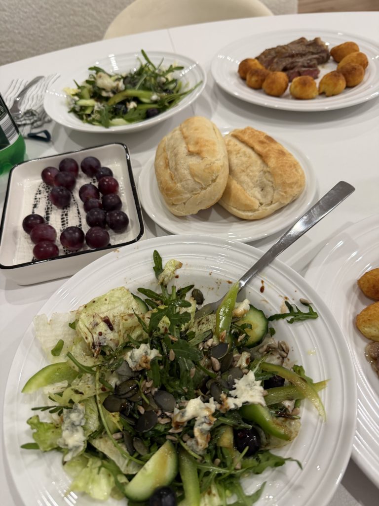 A dinner table with green salad, a bowl of grapes, two rolls, and a sliced steak surrounded by golden potato drops from Aldi