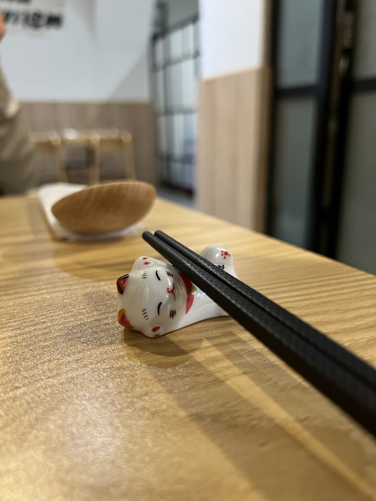 Chopsticks resting on ceramic cat chopstick rest, a wooden ramen spoon an Japanese decor softly in the background at Yuki Ramen Restaurant in Lisbon, Portugal