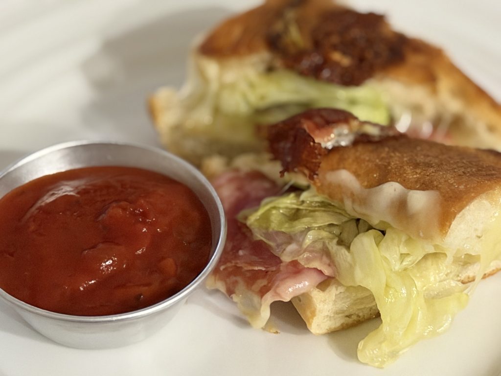 Cucina Arrabbiata Sauce Review — Close up of toasted Italian sandwich with Spianata Romana Roman Salami Mozarella Cheese and iceberg lettuce on a Baguette with a cup of sauce at the side  | Aldi Finds Portugal 